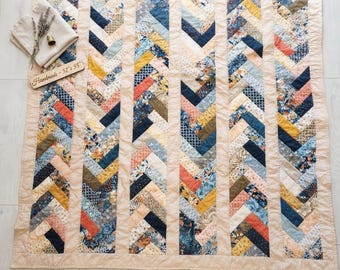 May include: A quilt featuring a herringbone pattern with vertical strips of fabric in shades of blue, yellow, orange, and cream. The quilt has a solid cream border and a wooden tag labelled "Handmade - 52" x 55"" resting on the corner.