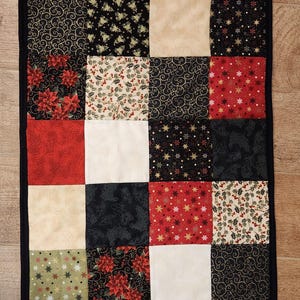 May include: A rectangular patchwork table runner featuring a grid of fabric squares in red, black, cream, and olive green. The fabric patterns include poinsettias, holly berries, gold swirls, and snowflakes, all bordered by a solid black fabric trim.