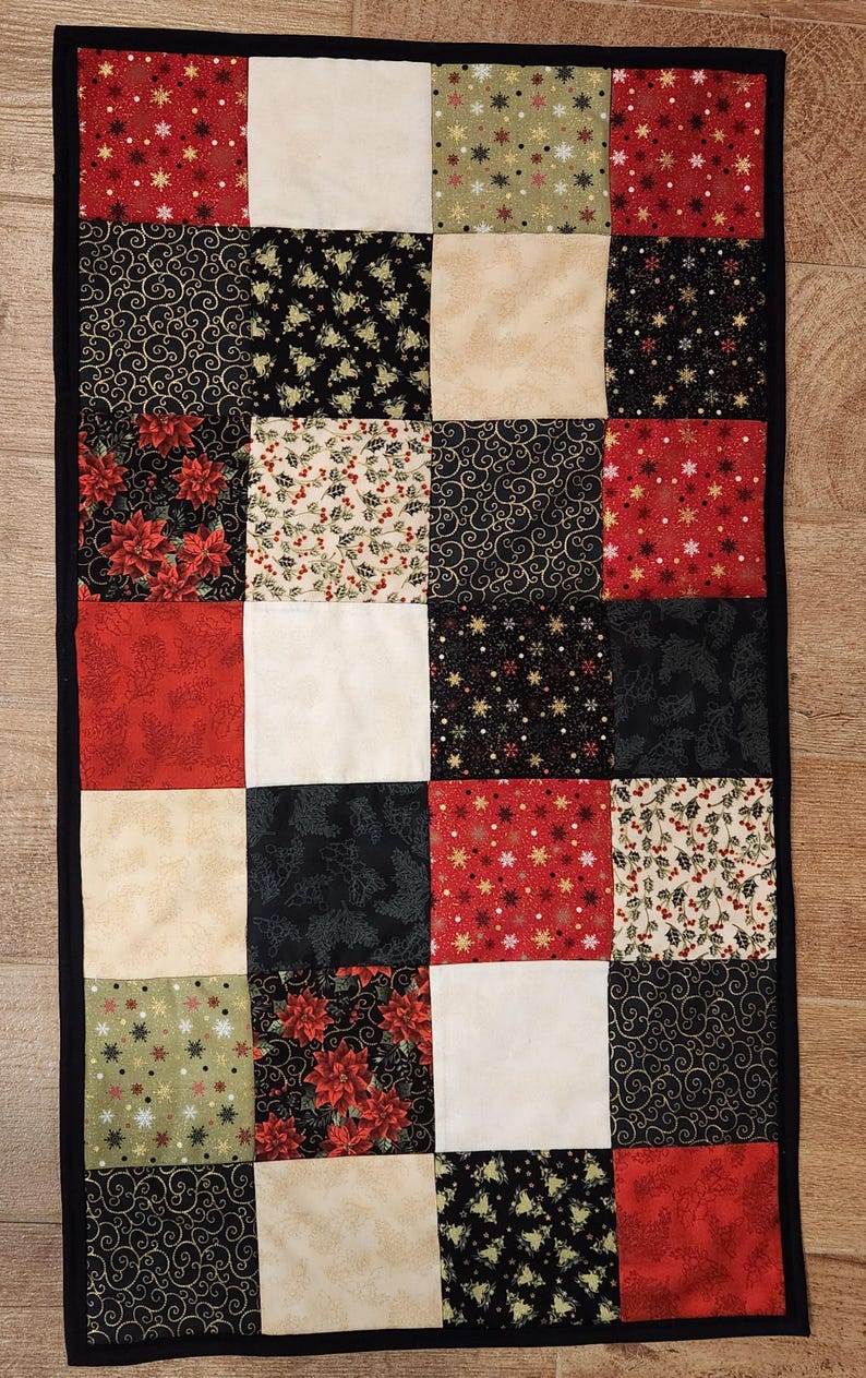 May include: A rectangular patchwork table runner featuring a grid of fabric squares in red, black, cream, and olive green. The fabric patterns include poinsettias, holly berries, gold swirls, and snowflakes, all bordered by a solid black fabric trim.