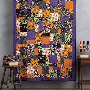 May include: A rectangular patchwork quilt featuring a variety of Halloween-themed fabric squares in shades of orange, purple, black, and lime green. The quilt is bordered in solid purple fabric and displays a repeating pattern of ghosts, pumpkins, and stars.