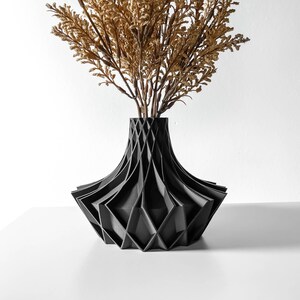 May include: A black geometric vase with a wide base and a narrow neck, holding dried, golden-brown decorative branches. The vase has a unique, angular design, and is set against a white background.