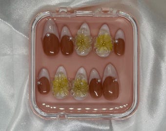 May include: A set of artificial nails in a clear, pink case. The nails feature a brown base with white tips and small pearl accents. Some nails have yellow flower embellishments. The case sits on a white, satin-like surface.