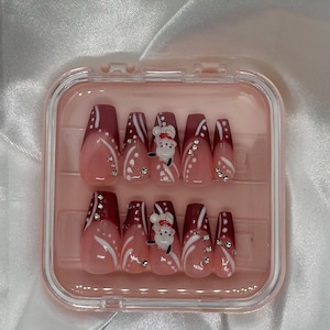 May include: A set of press-on nails in a clear plastic case. The nails feature a gradient design with a light pink base that fades into a deep burgundy. White accents and small rhinestones add detail, with a Santa Claus design on some nails.