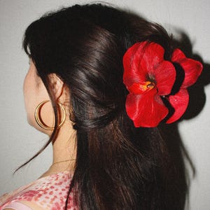 May include: A large, vibrant red orchid hair clip secured in dark brown hair. The accessory features realistic, layered petals with a deep crimson hue and a yellow center. A simple gold hoop earring is visible on the ear, and a pink patterned top is worn.