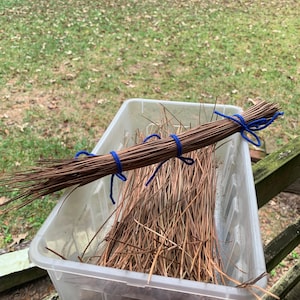 May include: A clear plastic container filled with brown pine needles. A bundle of pine needles is tied with blue yarn. The container sits on a wooden surface, with a blurred background of green grass and trees.