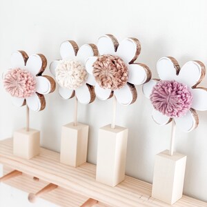 May include: Four white wooden flower shapes with brown centers and pink, beige, and brown pom-poms on a wooden shelf with hooks.