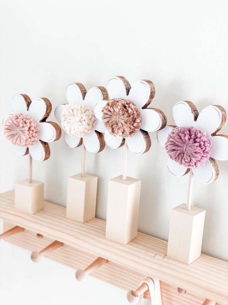 May include: Four white wooden flower shapes with brown centers and pink, beige, and brown pom-poms on a wooden shelf with hooks.