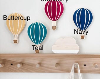 May include: Four wooden hot air balloon hooks in teal, navy, cranberry, and buttercup colors. The hooks are mounted on a wooden shelf with a white cloud shape below. The shelf has five wooden pegs.