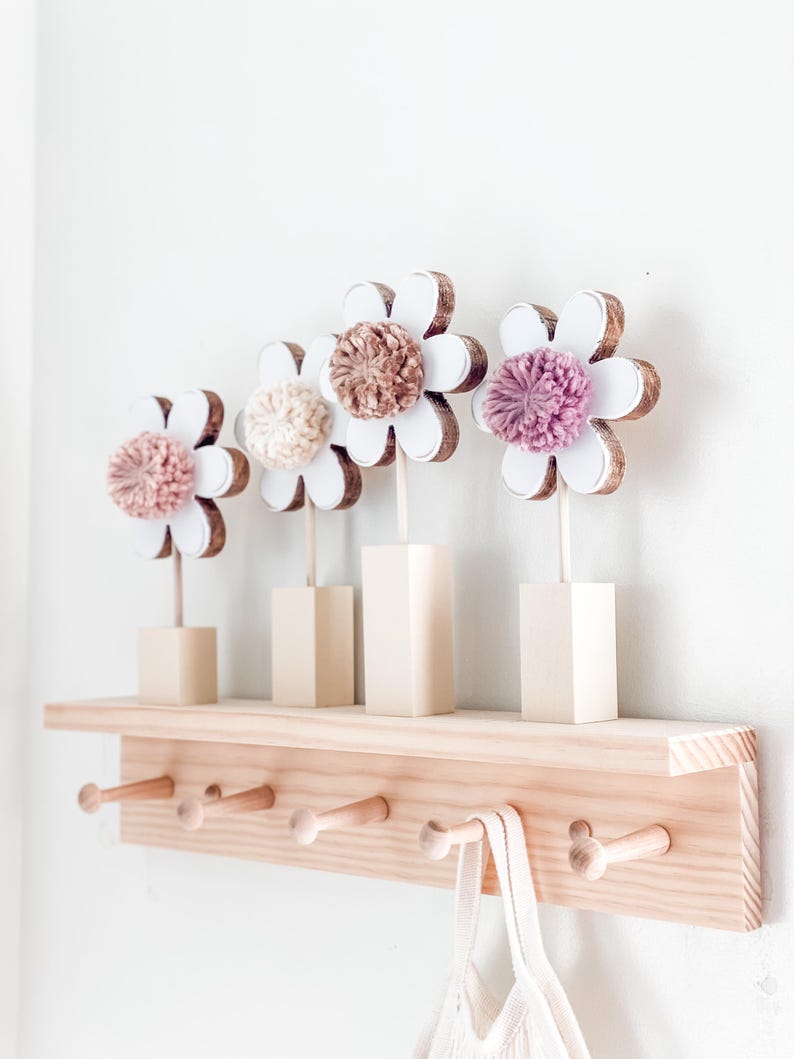 May include: A wooden shelf with five hooks and a white crocheted bag hanging on one of the hooks. Four wooden flower decorations with pink, brown, and white pom-poms are displayed on the shelf.