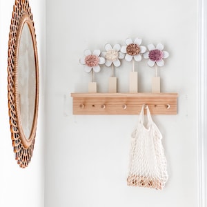 May include: A wooden shelf with five hooks and a white string bag hanging from one of the hooks. The shelf has four white flowers with pink, brown, and peach centers on top.