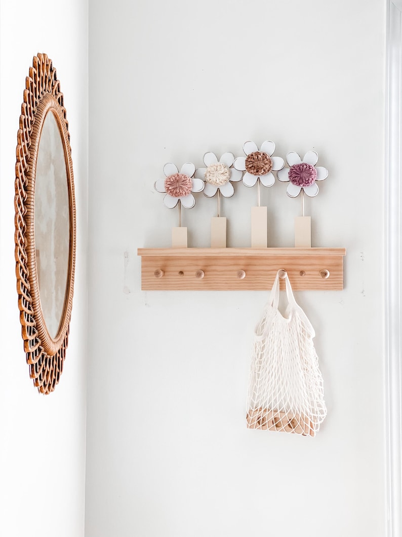 May include: A wooden shelf with five hooks and a white string bag hanging from one of the hooks. The shelf has four white flowers with pink, brown, and peach centers on top.