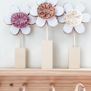 May include: Three wooden flower decorations with white painted petals and yarn centers. The flowers are in shades of pink, brown, and cream. The flowers are displayed on a wooden shelf with hooks.