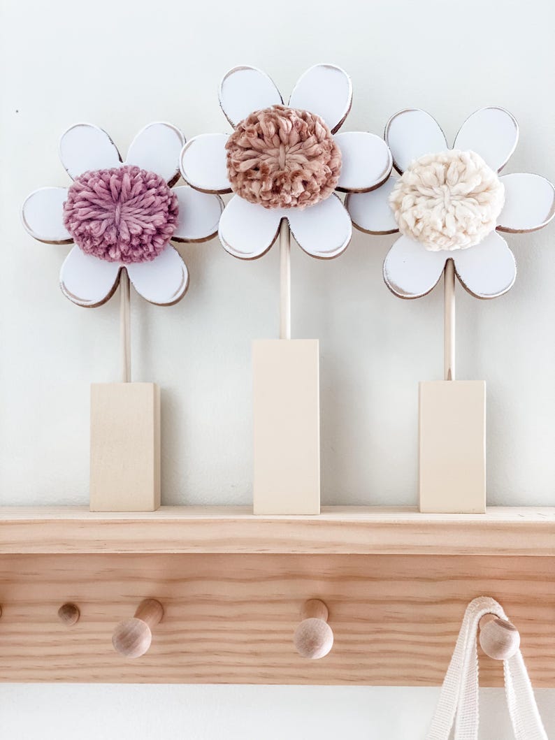 May include: Three wooden flower decorations with white painted petals and yarn centers. The flowers are in shades of pink, brown, and cream. The flowers are displayed on a wooden shelf with hooks.