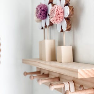 May include: A wooden shelf with five hooks and three wooden flower decorations with pink and purple pom-poms.