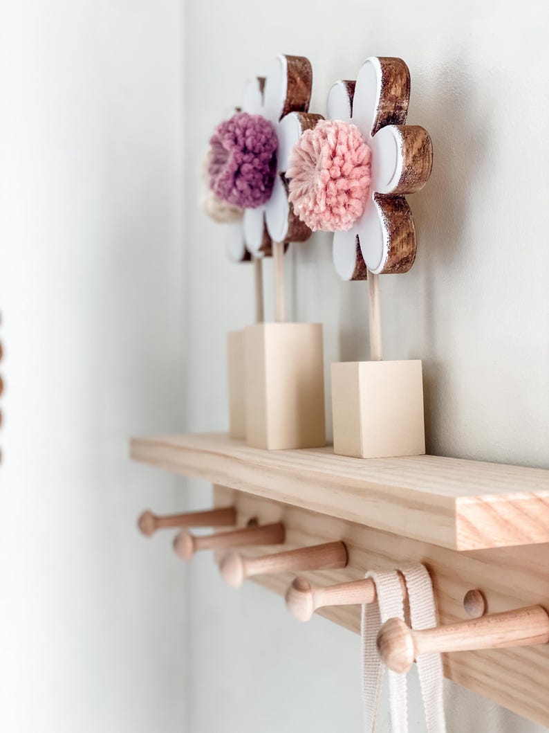 May include: A wooden shelf with five hooks and three wooden flower decorations with pink and purple pom-poms.