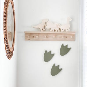 May include: A wooden shelf with five hooks and a row of wooden dinosaur cutouts. Three green dinosaur footprints are on the wall below the shelf.