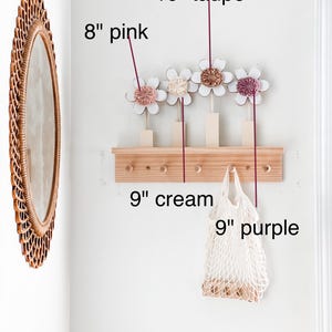 May include: A wooden wall-mounted rack with five hooks, decorated with four 8-inch pink, cream, and taupe felt flowers. A 9-inch cream-colored mesh bag hangs from one of the hooks.