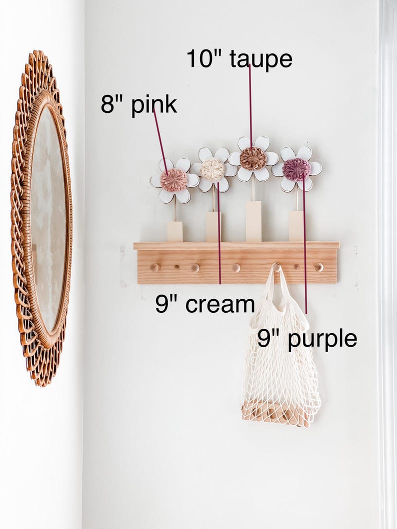 May include: A wooden wall-mounted rack with five hooks, decorated with four 8-inch pink, cream, and taupe felt flowers. A 9-inch cream-colored mesh bag hangs from one of the hooks.