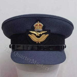 Puede incluir: Un sombrero de estilo militar azul oscuro con una banda negra y un emblema bordado en oro que representa una corona y un pájaro con las alas extendidas. El emblema está centrado en la parte delantera del sombrero.