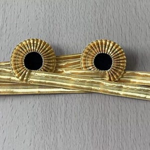 Puede incluir: Dos adornos circulares de color dorado con centros negros, unidos a una cinta dorada. Los adornos tienen un diseño radial.
