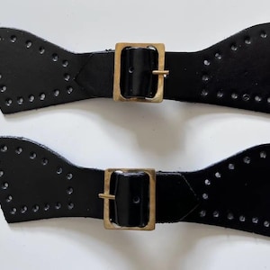 Puede incluir: Dos correas de cuero negro con hebillas doradas. Las correas tienen un diseño decorativo con agujeros alrededor de los bordes.
