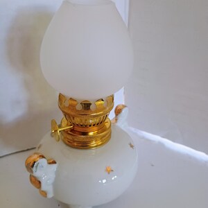 Peut inclure: Une lampe à huile en céramique blanche avec des détails dorés et un abat-jour en verre dépoli. La lampe a une base ronde avec trois petits pieds et des motifs d'étoiles. Le dessus et les détails sont dorés.