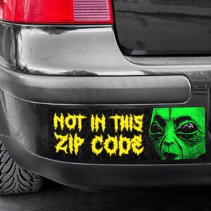 Peut inclure: Un pare-chocs de voiture noir avec un autocollant jaune et noir qui indique "Pas dans ce code postal". L'autocollant présente également un visage d'extraterrestre vert.