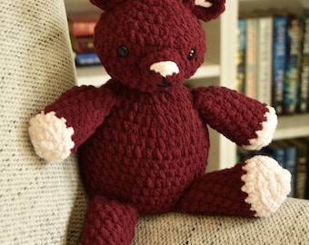Può includere: Un orsacchiotto all'uncinetto bordeaux fatto a mano con dettagli bianchi sulle zampe, orecchie e muso. Il peluche è seduto su una sedia grigia imbottita. L'orso ha occhi neri a bottone e un piccolo sorriso nero cucito.