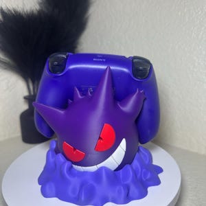 Op de afbeelding: Een paarse controllerstandaard met een 3D-geprint Gengar-ontwerp met rode ogen en een brede grijns. De standaard is ontworpen om een paarse Sony PlayStation-controller te bevatten. De standaard staat op een wit oppervlak.