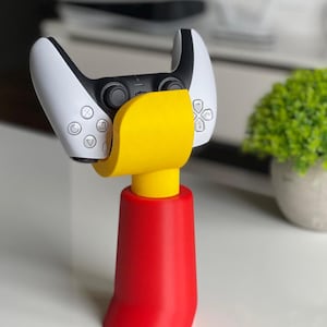 May include: A yellow and red 3D printed stand for a white and black video game controller. The stand is shaped like a stylised figure with a rounded base.