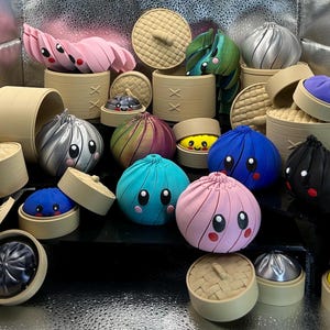 May include: A collection of small, colorful, round dumpling figurines with expressive faces, each featuring large black eyes and pink cheeks. The figures are rendered in various solid colors including pink, blue, silver, purple, and black, and are displayed with tan, cylindrical, woven-textured steamer baskets.