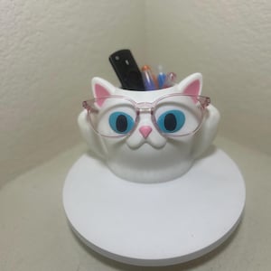 May include: A white cat-shaped desk organizer with large blue eyes, a pink nose, and pink inner ears. The cat figure wears translucent pink-rimmed glasses and holds several pens and a black remote control inside its hollow top opening.