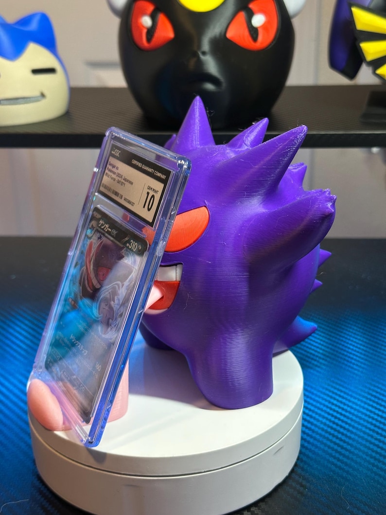 May include: A purple and red Pokemon card holder featuring a 3D printed Gengar figure holding a graded Pokemon card in a clear plastic case. The card is graded a 10 by the Certified Guaranty Company.