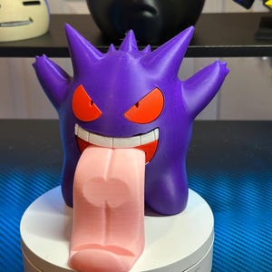 May include: A purple and pink 3D printed figurine of a Pokemon character,  Gengar, with a large, pink tongue sticking out. The figurine is on a white base.