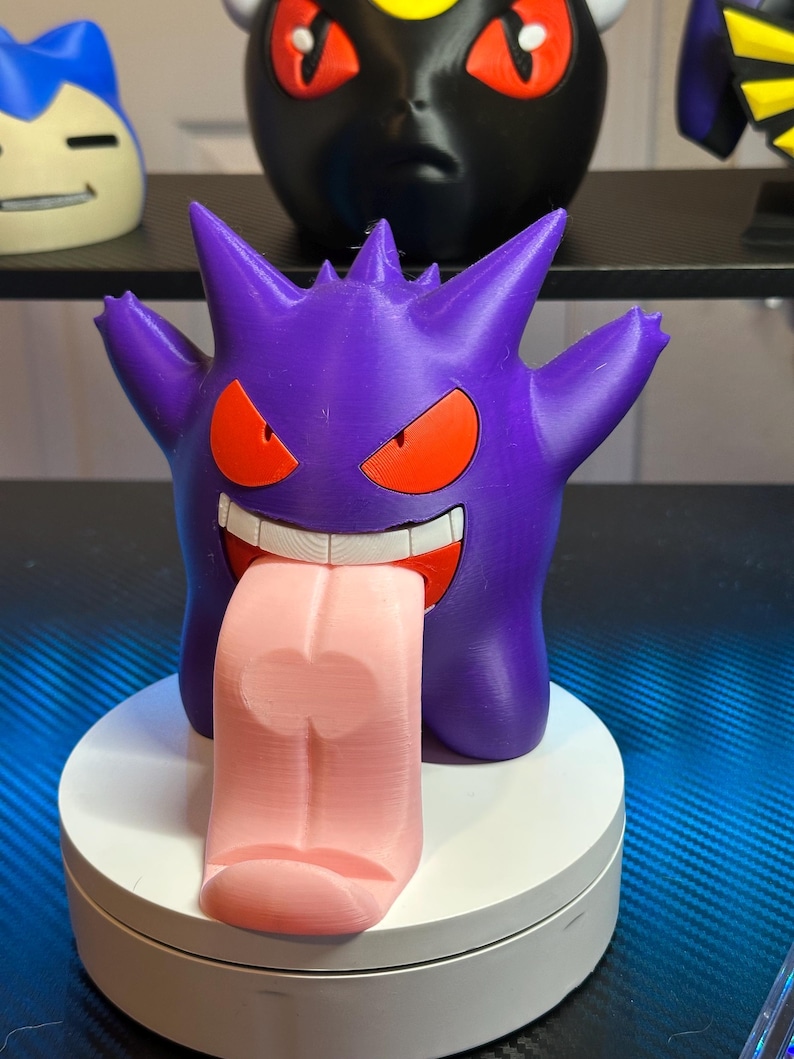 May include: A purple and pink 3D printed figurine of a Pokemon character,  Gengar, with a large, pink tongue sticking out. The figurine is on a white base.