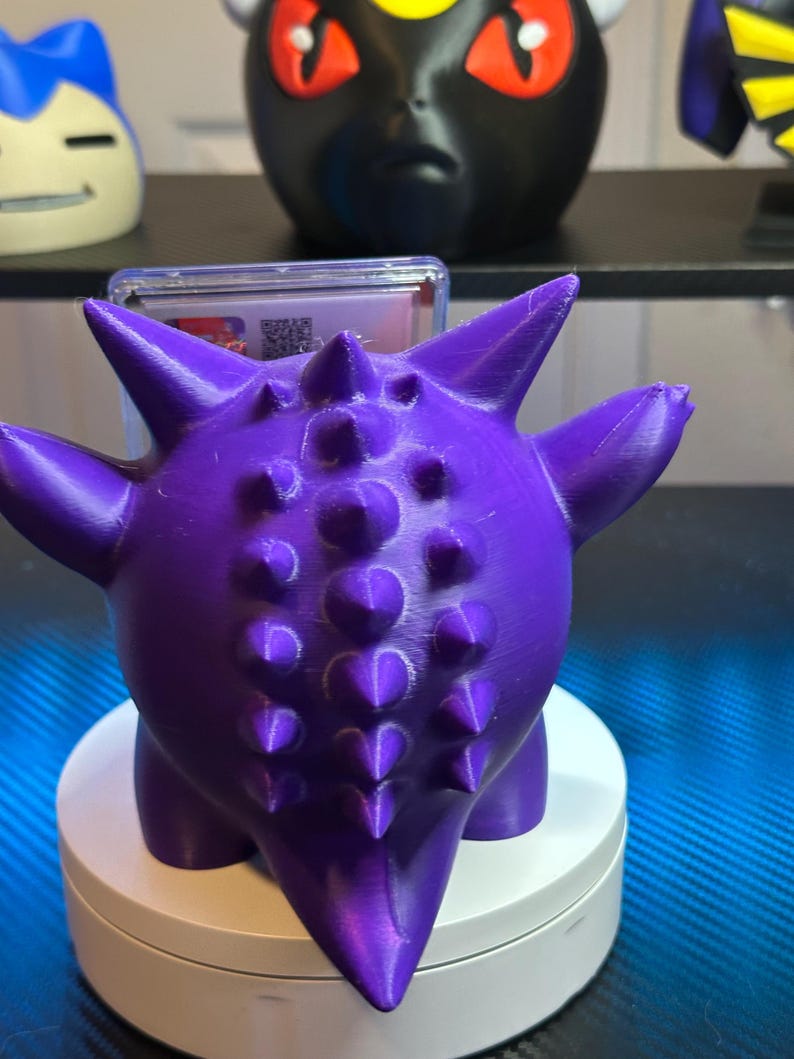 May include: A purple 3D printed Pokemon figure of a Spinarak with a white base. The figure has a spiky back and is holding a clear plastic card.