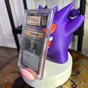 May include: A purple and red Pokemon card holder with a clear plastic case holding a Japanese Pokemon Trainer card. The card features a character named "Oak" and the text "Trainer" and "Support". The card holder is on a white base.