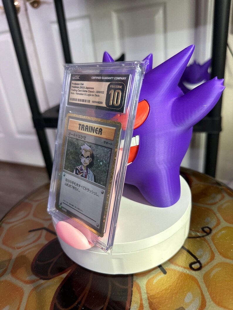 May include: A purple and red Pokemon card holder with a clear plastic case holding a Japanese Pokemon Trainer card. The card features a character named "Oak" and the text "Trainer" and "Support". The card holder is on a white base.
