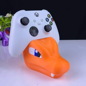May include: A white Xbox controller is resting in a bright orange, cartoon-style stand shaped like a Pokemon character. The stand has a blue eye and sharp teeth.