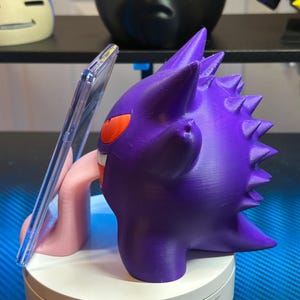 May include: A purple and pink 3D printed Gengar phone stand with a clear plastic phone holder. The Gengar is holding the phone holder in its mouth and has a long pink tongue sticking out.