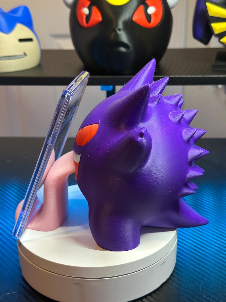 May include: A purple and pink 3D printed Gengar phone stand with a clear plastic phone holder. The Gengar is holding the phone holder in its mouth and has a long pink tongue sticking out.