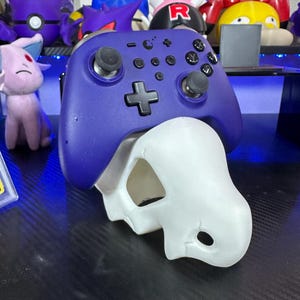 May include: A purple video game controller rests atop a white, skull-shaped object. The controller has black buttons and joysticks. Plush toys of various colors and shapes are visible in the background, suggesting a gaming or collectible theme.