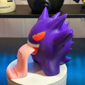 May include: A purple and pink 3D printed figurine of a Pokemon character with a large tongue sticking out. The figurine is on a white base.