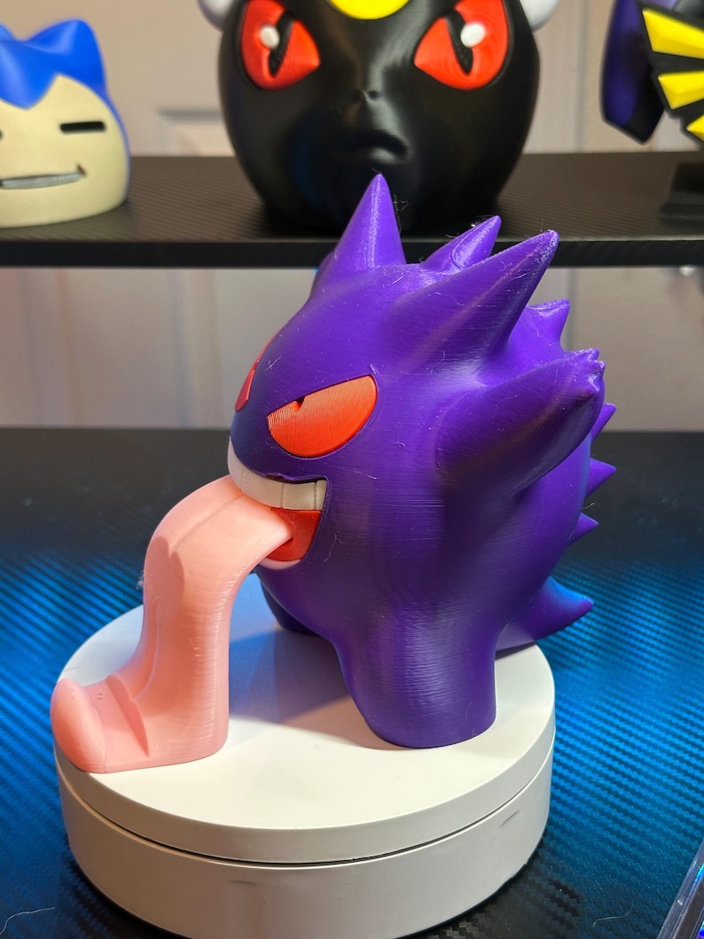 May include: A purple and pink 3D printed figurine of a Pokemon character with a large tongue sticking out. The figurine is on a white base.