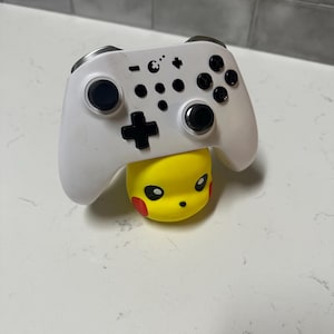 Könnte beinhalten: Ein weißer Gamecontroller mit schwarzen Knöpfen liegt auf einer gelben Pikachu-Figur. Der Controller hat ein symmetrisches Design mit zwei Analogsticks und verschiedenen Tasten. Die Pikachu-Figur hat rote Wangen und einen lächelnden Gesichtsausdruck.