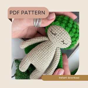 May include: A crochet pattern for a small, beige doll with a green crocheted hat. The doll has closed eyes and a sweet expression. The pattern is available for instant download.