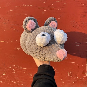 May include: A gray crocheted mouse head with pink inner ears and nose, and white eyes. The handmade plushie is held up against a red background. The mouse head is a whimsical, soft toy.