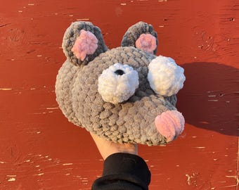 May include: A gray crocheted mouse head with pink inner ears and nose, and white eyes. The handmade plushie is held up against a red background. The mouse head is a whimsical, soft toy.