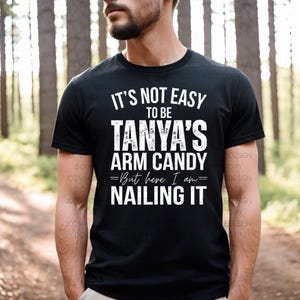 May include: Black t-shirt with white text that reads "IT'S NOT EASY TO BE TANYA'S ARM CANDY =But here I am= NAILING IT". The shirt is a crew neck style.