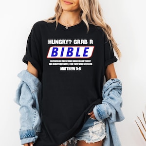 Puede incluir: Camiseta negra con el texto "HUNGRY? GRAB A BIBLE" en blanco y azul. Debajo del título está el texto "BLESSED ARE THOSE WHO HUNGER AND THIRST FOR RIGHTEOUSNESS, FOR THEY WILL BE FILLED MATTHEW 5:6".